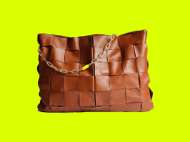 Bolsa feminina de couro na cor marrom, com cortes quadrados, e alça dourada em formato de corrente com detalhe verde, em fundo verde limão