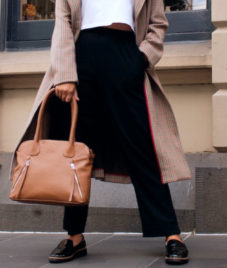 Cintura, pernas e pés de modelo feminina vestindo sobretudo de cor predominante bege com detalhes em tom vermelho e escuro, com calça e par de sapatos na cor preta, segurando bolsa na cor bege, com fundo de prédio
