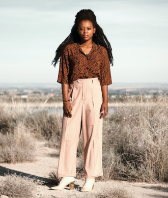 Modelo feminina vestindo blusa marrom, calça bege, par de sapatos altos na cor branca, cordão e pulseira, em um ambiente aberto com chão de terra e matos secos