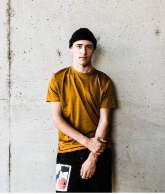 Modelo masculo vestindo touca preta, blusa marrom e calça preta, com uma parede cinza de fundo
